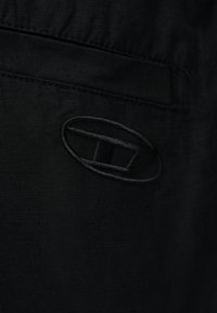 Μαύρο ύφασμα με κεντημένο λογότυπο που απεικονίζει ένα οβάλ σχήμα που περιβάλλει ένα σχηματοποιημένο «T» κοντά σε μια οριζόντια ραφή ή τσέπη.