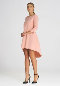 Pfirsichfarbenes Kleid mit asymmetrischem Saum, Dreiviertelärmel, aus glattem Stoff. Kombiniert mit schwarzen Stilettos.