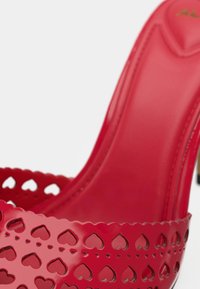 Sandale à talons hauts rouge en matériau brillant, présentant des motifs de cœurs découpés le long de la bride et une tige lisse et moulée.