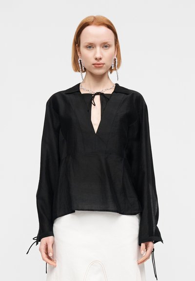 Blouse en lin noir avec un décolleté en V profond, manches longues et liens ajustables aux poignets. Le modèle présente une coupe ample et des coutures sur le panneau avant.