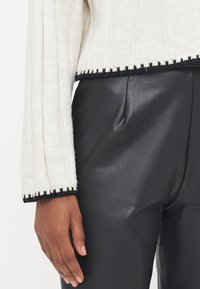 Knit sweater in off-white with ribbed texture and black trim, paired with smooth black leather pants. Hand visible at the edge.