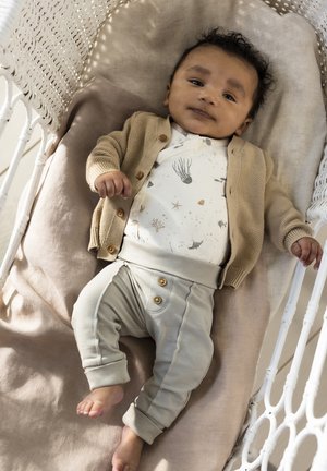 Lichtbeige gebreide cardigan over een gebloemd rompertje met maritieme motifs; grijze broek met knopen; rustend op een zachte taupe deken in een geweven wieg.