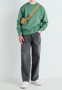 Sudadera verde con cuello redondo, combinada con jeans grises de corte holgado. Se lleva una bolsa cruzada color topo y unas zapatillas blancas completan el look.