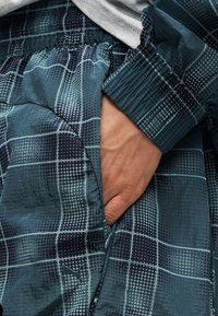 Χέρι μέσα στην τσέπη με φερμουάρ ενός σκούρου μπλε, εμπριμέ μπουφάν με ελαστικό μανίκι και διακριτικό λογότυπο Puma.