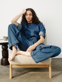 Moteris su ilgais garbanotais plaukais sėdi basomis ant smėlio spalvos kėdės, vilkėdama suknele su trumpomis kelnėmis, atsiremdama galva į ranką, šalia mažo juodo šoninio stalo.