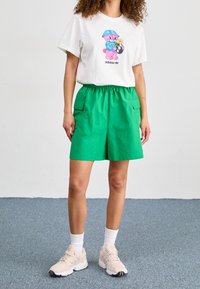 Camiseta blanca con gráfico de oso colorido y logo, combinada con pantalones cortos verdes a la altura de la rodilla con bolsillos cargo. Zapatillas ligeras y calcetines blancos.