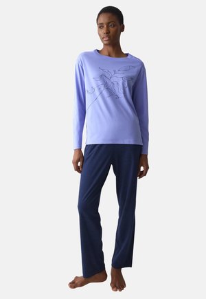 Femme debout pieds nus portant un haut lavande à manches longues avec un motif floral noir et un pantalon bleu marine sur un fond clair.