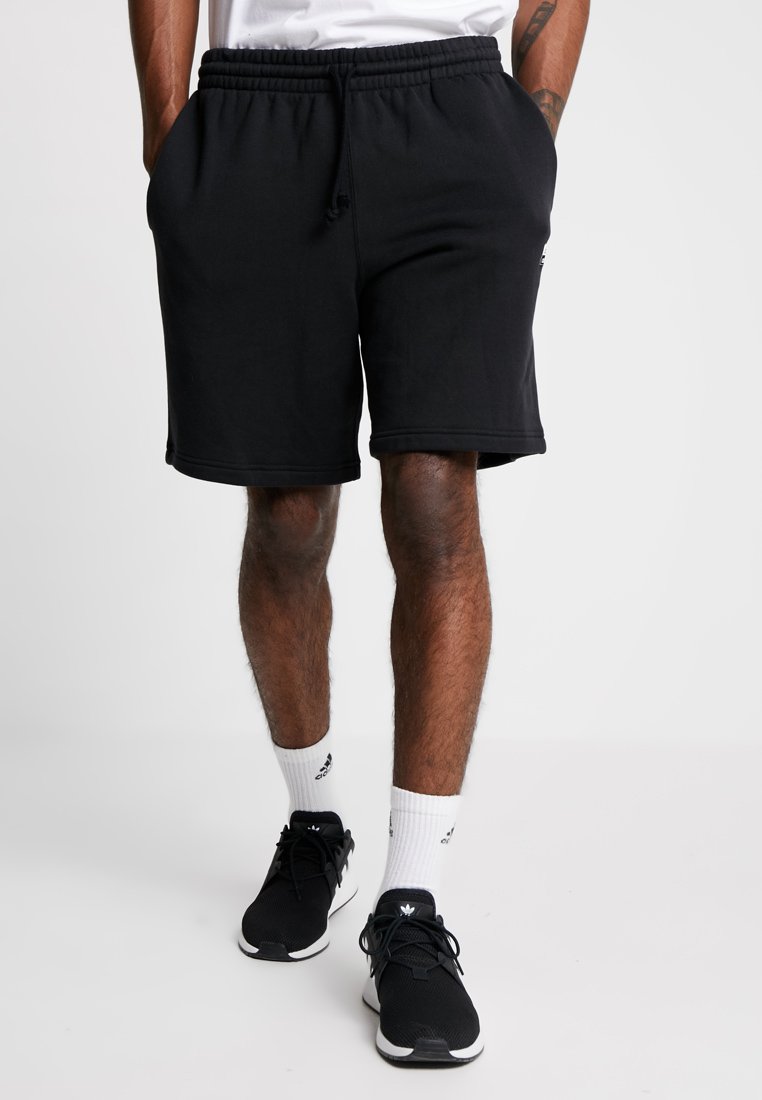 Pantaloni corti in cotone nero con vita elastica e cordoncino, dotati di tasche laterali e un taglio rilassato, abbinati a sneakers nere e calzini bianchi.