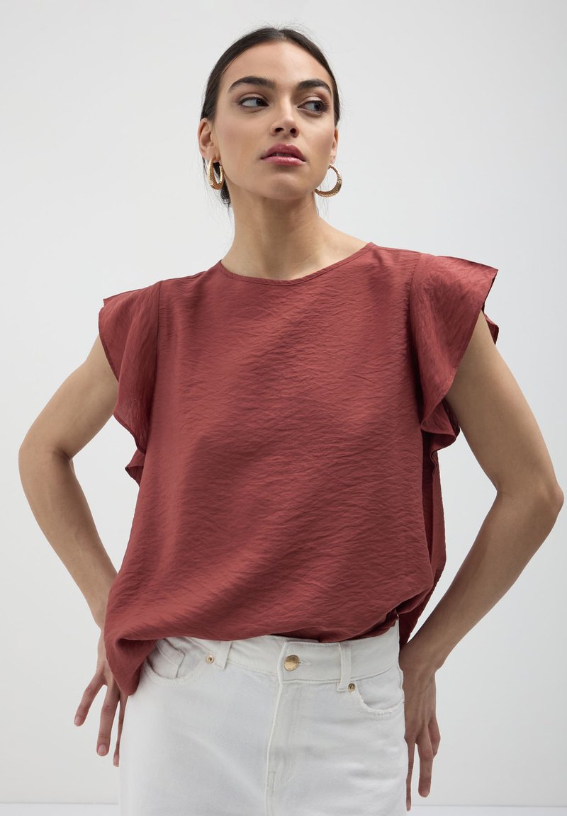 Donna che indossa un top smanicato color ruggine con spalline arricciate e jeans bianchi a vita alta, che guarda di lato con le mani sui fianchi.