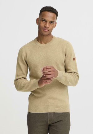 Light brown knit sweater with a round neckline, ribbed cuffs, and hem. Features a small brown logo patch on the left sleeve.