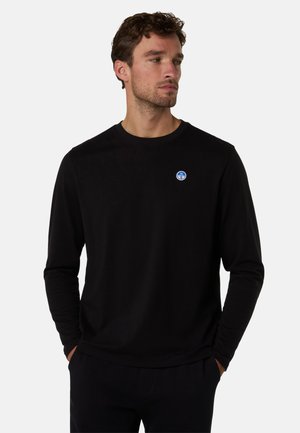 Uomo che indossa una maglia nera a maniche lunghe con un piccolo logo circolare blu e bianco sul petto, mani nelle tasche, guarda di lato.