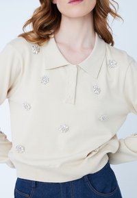 Pullover lavorato a maglia beige con colletto classico, caratterizzato da decorazioni floreali in tonalità argentata sparse sul davanti e sulle maniche.