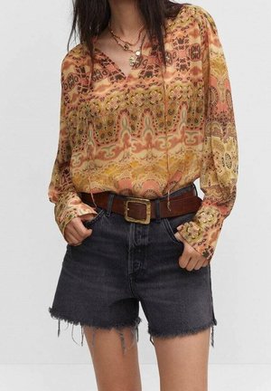 Langärmlige, gemusterte Bluse in warmen Orangetönen und Gelbtönen, kombiniert mit dunklen Denim-Shorts. Mit fransigen Kanten und einem braunen Gürtel.