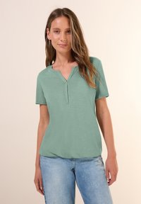 Haut vert menthe à manches courtes avec un décolleté en V et une coupe décontractée. Texture douce et légère brillance. Associé à un jean bleu clair.