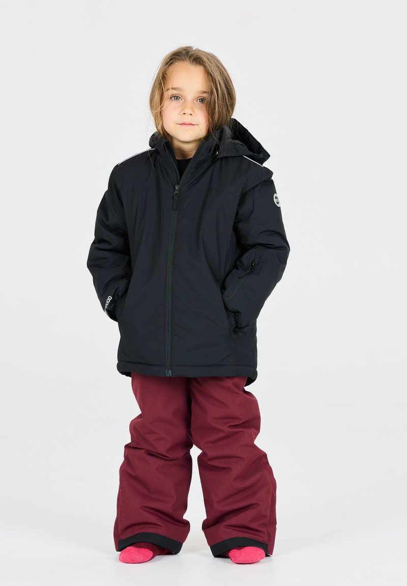 Schwarze isolierte Jacke mit Kapuze, Reißverschluss und seitlichen Taschen. Burgunderrote Schneehosen, lockere Passform und kontrastierende pinke Socken.
