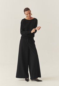 Schwarzer Pullover mit strukturierten Nietenakzenten, weit geschnittene schwarze Hose und spitze schwarze Absätze. Minimalistisches Design mit entspannter Passform und Rundhalsausschnitt.