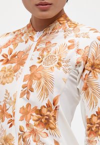 Floral zip-up rash guard featuring orange and brown tropical patterns on a white base. Soft, stretchy fabric with flat seams and logo detail.