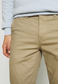 Pantaloni beige in cotone con finitura texturizzata, dotati di tasche anteriori e sagoma aderente. In vita è visibile una felpa azzurro chiaro.