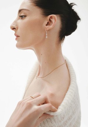 Mujer con suéter de punto blanco sin hombros, con el cabello oscuro recogido en un moño, usando pendientes delicados, un collar delgado y un anillo, mirando hacia la izquierda de perfil.