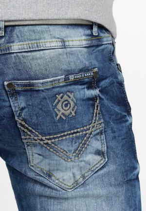 Un jean en denim bleu présente un délavage, une poche arrière avec un motif brodé, et des coutures contrastées. Design de poche distinct avec des coutures décoratives.