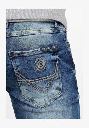Blauwe denim jeans hebben een vervaagde wassing, een achterzak met geborduurd detail en contrasterende stiksels. Opvallend zakontwerp met decoratieve stiksels.