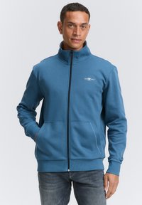 Blå zip-up sweatshirt lavet af blødt stof, med høj krave, to forlommer og et lille hvidt logo på brystet.