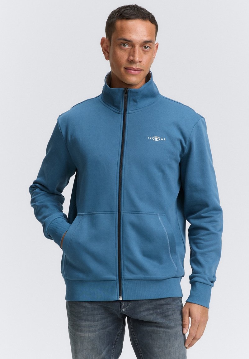 Blå zip-up sweatshirt lavet af blødt stof, med høj krave, to forlommer og et lille hvidt logo på brystet.