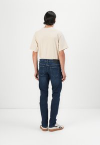 Camiseta de manga corta beige combinada con unos vaqueros ajustados azul oscuro. Los vaqueros tienen un parche de cuero en la parte trasera y dos bolsillos traseros. Zapatillas blancas.