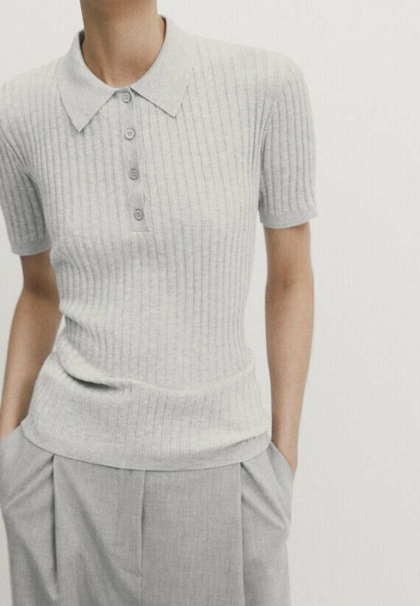 Personne portant un polo gris clair à manches courtes côtelé, rentré dans un pantalon plissé gris taille haute, les mains dans les poches.
