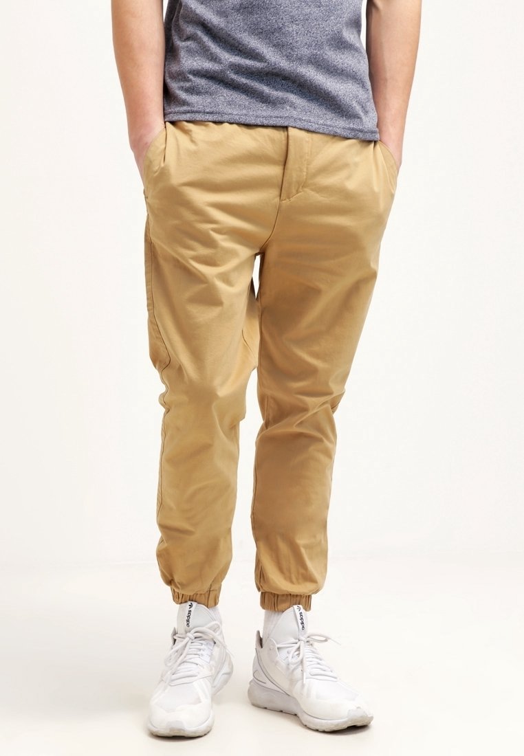 Osoba ubrana w beżowe spodnie dresowe z elastycznymi mankietami, szary heatherowy T-shirt, białe skarpetki i białe sneakersy Adidas, stojąca na białym tle.