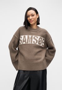 Camisola castanha em malha com uma faixa branca com logótipo no peito, punhos e bainha canelados, e um corte descontraído. Usada com calças pretas de couro.