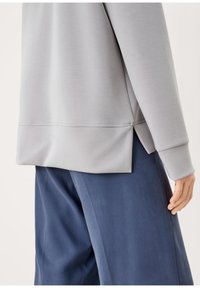 Sudadera gris claro con un ajuste relajado, que presenta una abertura lateral en el dobladillo. Combinada con pantalones azul marino, mostrando una textura suave.