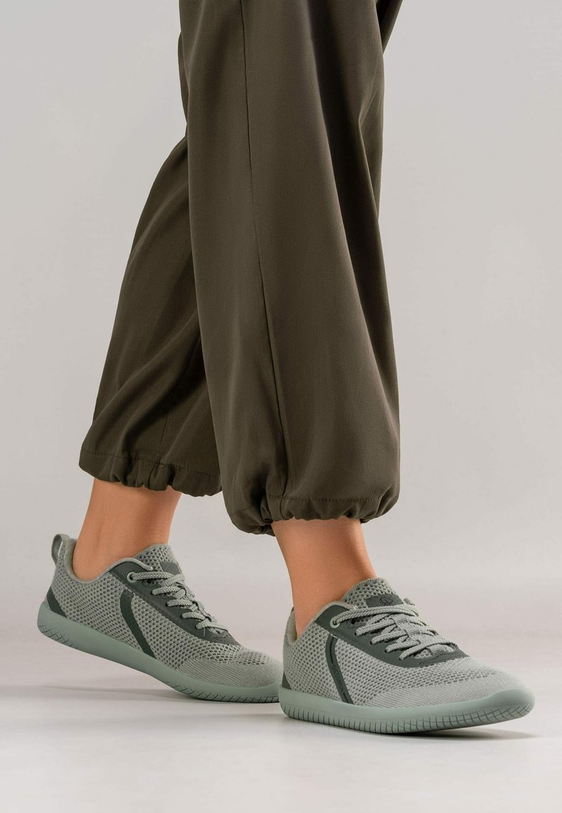 Pieds portant des baskets en maille respirante grise avec lacets, assorties à un pantalon vert olive retroussé, sur un fond clair uni.