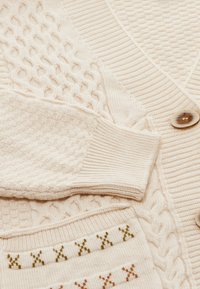 Maglioni lavorati a maglia di colore crema con vari motivi, polsini a coste e bottoni in primo piano.