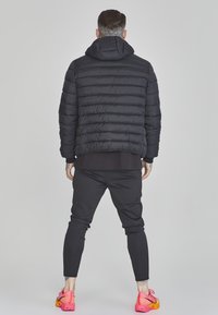 Veste matelassée noire avec capuche, présentant un matelassage horizontal. Portée avec un pantalon noir ajusté et des baskets colorées.