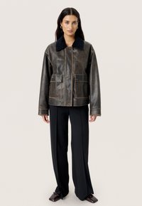 Brun læderjakke med en sort fløjelskraæg, der har to frontlommer, synlige syninger og en afslappet pasform. Bærer sorte bukser.