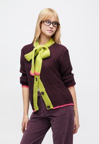 Megztinis tamsiai violetinės spalvos su laimo žalumos kaspinu ir rožiniais akcentais. Yra dideli mygtukai ir faktūruoto audinio detalė.