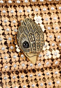 Złota kopertówka z metalowymi cekinami i kwiatowymi akcentami. Posiada teksturowany zamek w kształcie ptaka z czarnymi i złotymi detalami. Iskrzące ozdoby.