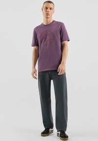 Fioletowa bawełniana koszulka z okrągłym nadrukiem „QUIET SENSE”. Noszona z luźnymi ciemnymi jeansami i czarnymi trampkami z białym akcentem.