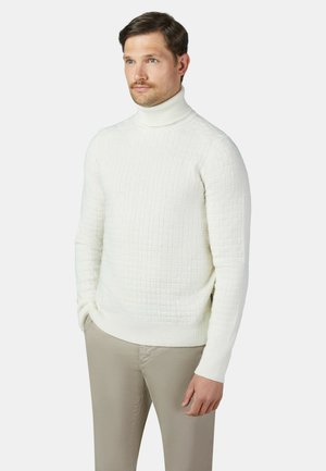 Uomo con capelli castano chiaro che indossa un maglione bianco a collo alto con trama e pantaloni beige, in piedi davanti a uno sfondo semplice.