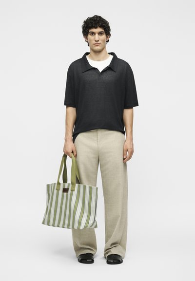 Νέος άνδρας φοράει μαύρο πουκάμισο με κοντά μανίκια και γιακά, μπεζ παντελόνι με φαρδιά μπατζάκια, μαύρα σανδάλια, κρατώντας μια πράσινη και λευκή ριγέ τσάντα.