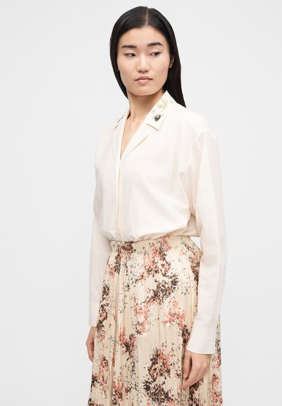 Young woman wearing a cream blouse with floral embroidery on the collar and a pleated skirt with pink and black floral patterns.