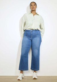 Heledad sinine denim laiade jalgadega teksad eespooltaskutega, seljas valge ja rohelise triibuline nööbita särk ning valged tossud.
