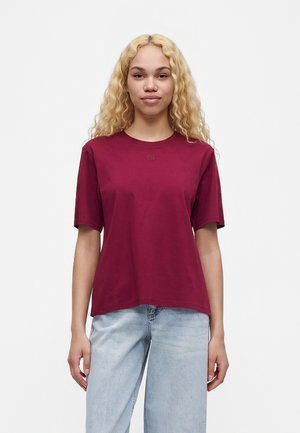 Jeune femme aux longs cheveux blonds bouclés portant un t-shirt bordeaux uni et un jean bleu clair, debout devant un fond blanc.
