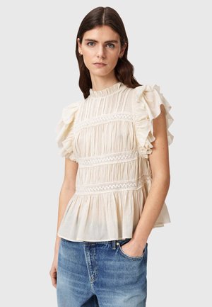 Kobieta ubrana w kremową, falbanianą bluzkę bez rękawów z koronkowymi detalami, połączoną z niebieskimi dżinsami, na jednolitym tle.