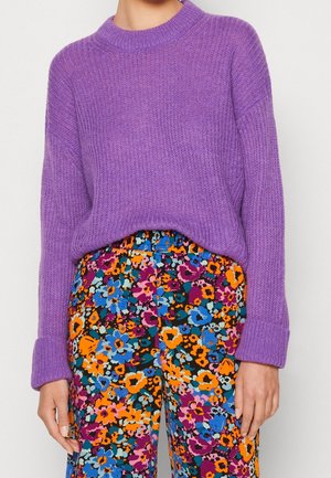 Kobieta w fioletowym swetrze dzianym, włożonym do wysokich spodni z jasnym, wielokolorowym kwiatowym wzorem.