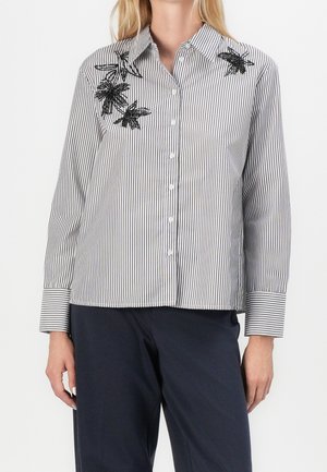 Femme portant une chemise blanche boutonnée avec de fines rayures noires et une broderie florale noire sur le côté supérieur gauche, associée à un pantalon foncé.