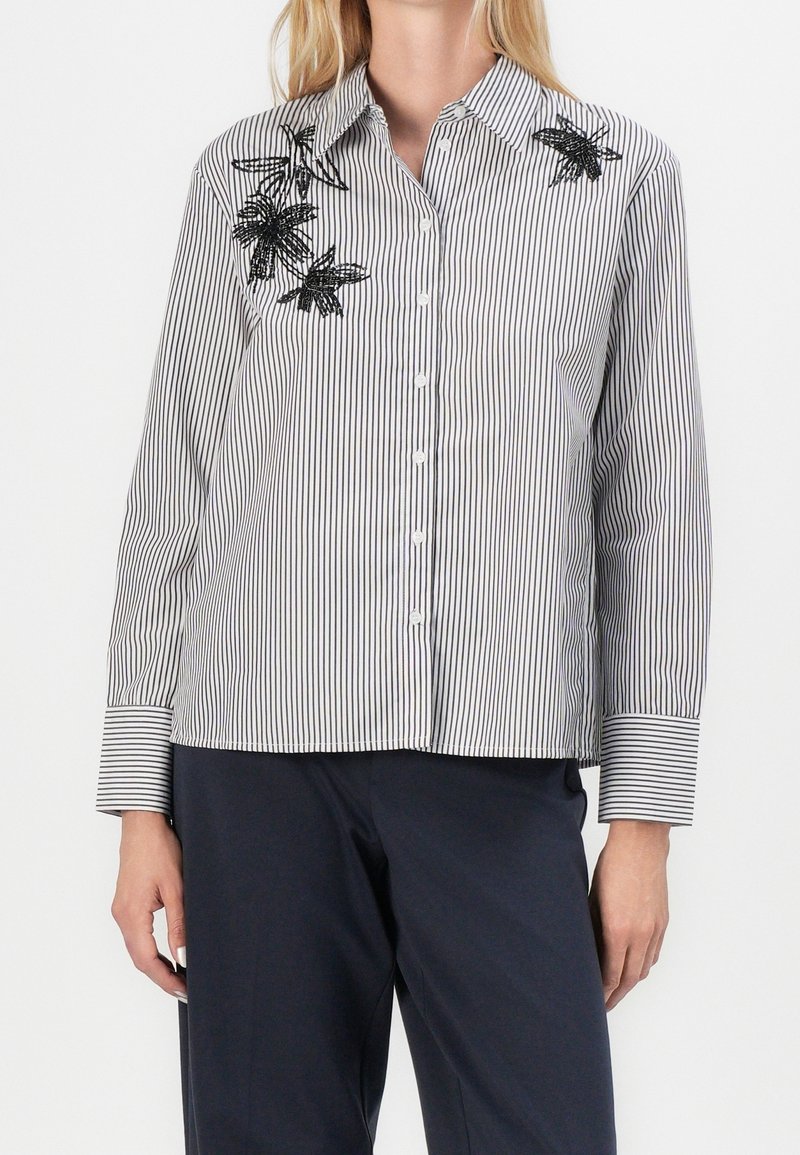 Femme portant une chemise blanche boutonnée avec de fines rayures noires et une broderie florale noire sur le côté supérieur gauche, associée à un pantalon foncé.