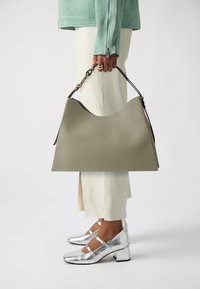 Borsa a tracolla verde in pelle con una distintiva forma a onda nella parte superiore, tenuta da una persona che indossa pantaloni beige e scarpe con tacco argentate.