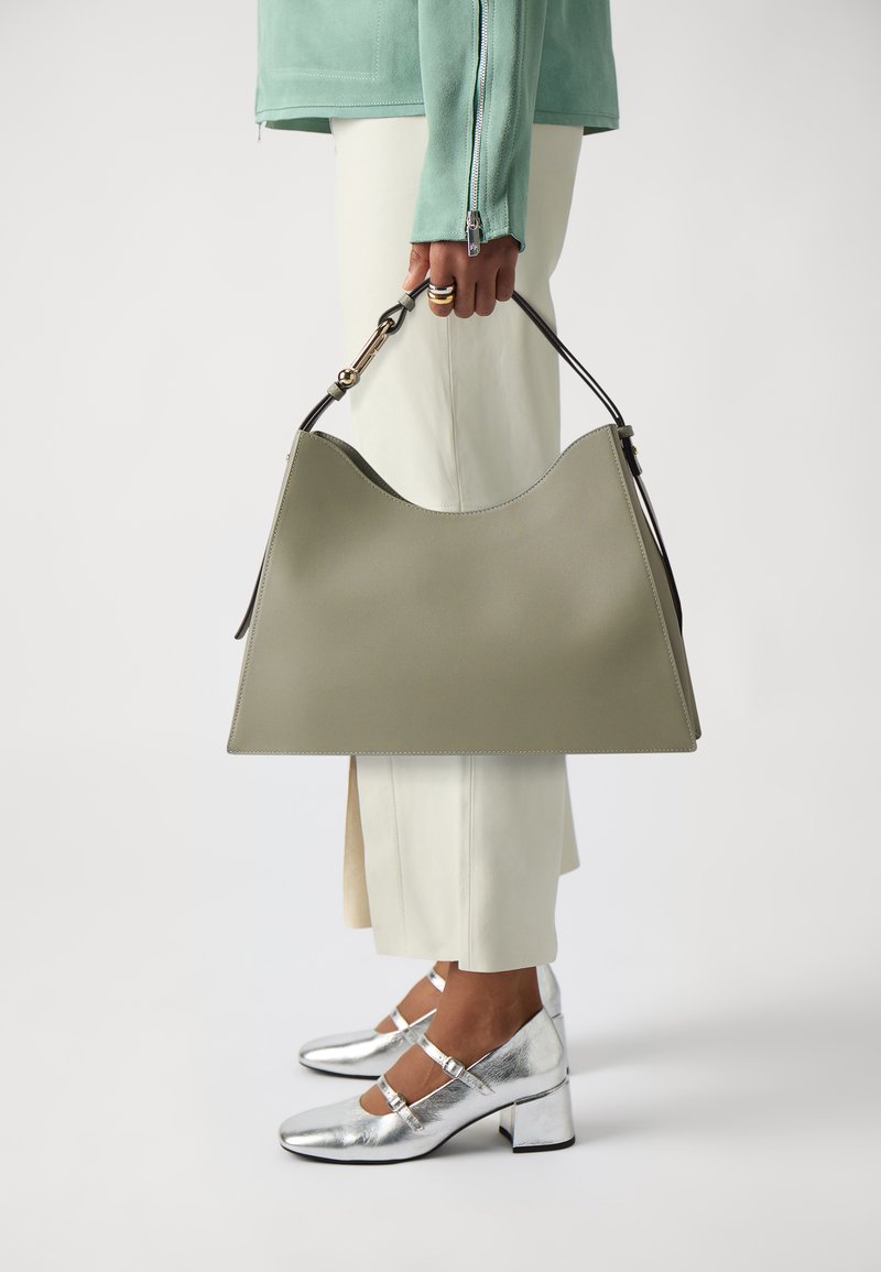 Borsa a tracolla verde in pelle con una distintiva forma a onda nella parte superiore, tenuta da una persona che indossa pantaloni beige e scarpe con tacco argentate.